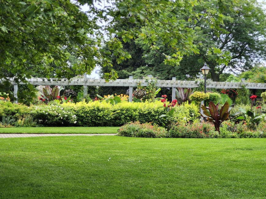 A well-maintained garden features a lush, evenly cut green lawn in the foreground, bordered by a paved pathway. Behind the lawn, there are vibrant flower beds and shrub borders with a variety of color