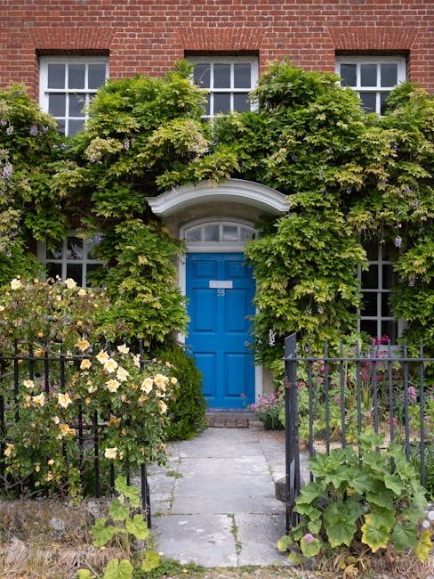 A charming front garden of a red-brick house featuring a bright blue front door with a semi-circular white arch above it. The entrance is framed by lush green foliage, including climbing plants with b